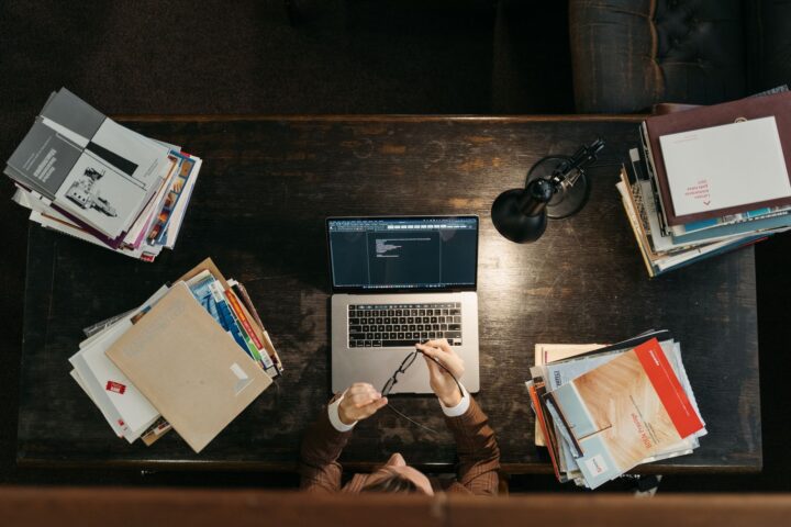 暗い木のデスクを上から見たところ。ノートパソコン、書類の束、ヘッドホンが並び、手元だけが写っている